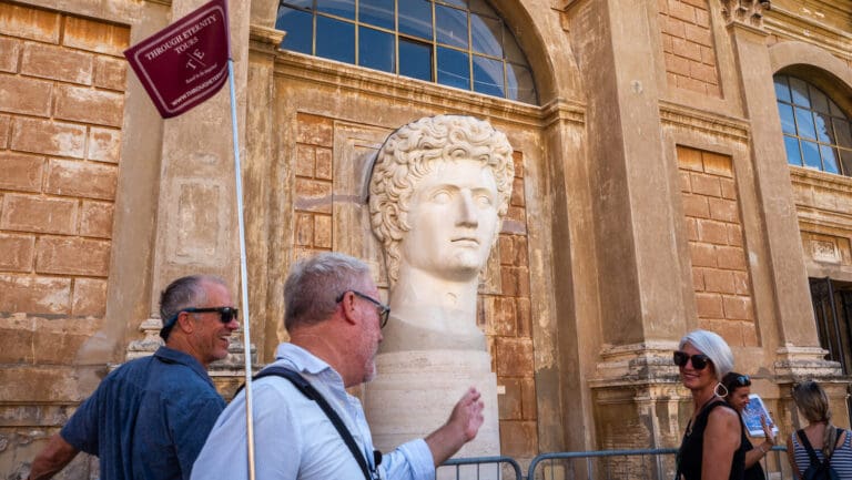 Tourists stroll past a Roman emperor statue in front of a historic building.
